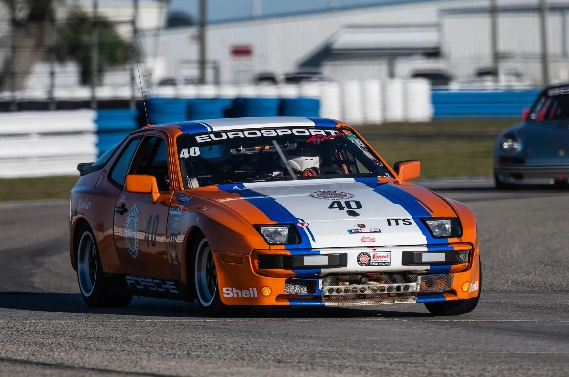 A vintage race car, painted in orange and white with blue stripes, driving on a race track, with a blurred background of other cars and track barriers.
