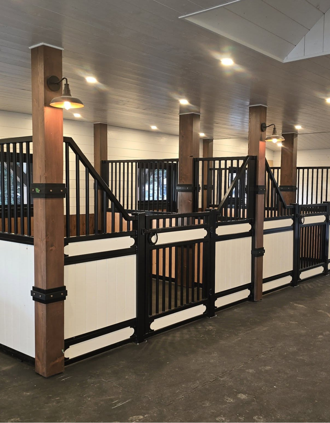 Interior of a stable with black metal railings, wooden posts, and white paneling, illuminated by ceiling lights.