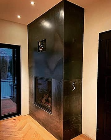 Modern fireplace with a black mantle and built-in television, located in a cozy living room with wooden flooring and a sliding glass door.