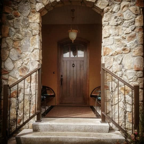 Stone archway leading to a wooden front door with black hardware, flanked by two metal handrails, with a chandelier hanging overhead.