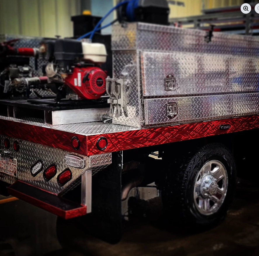 Close-up of the back of a fire truck with diamond plate metal compartments and a red stripe. Part of the rear wheel is visible.