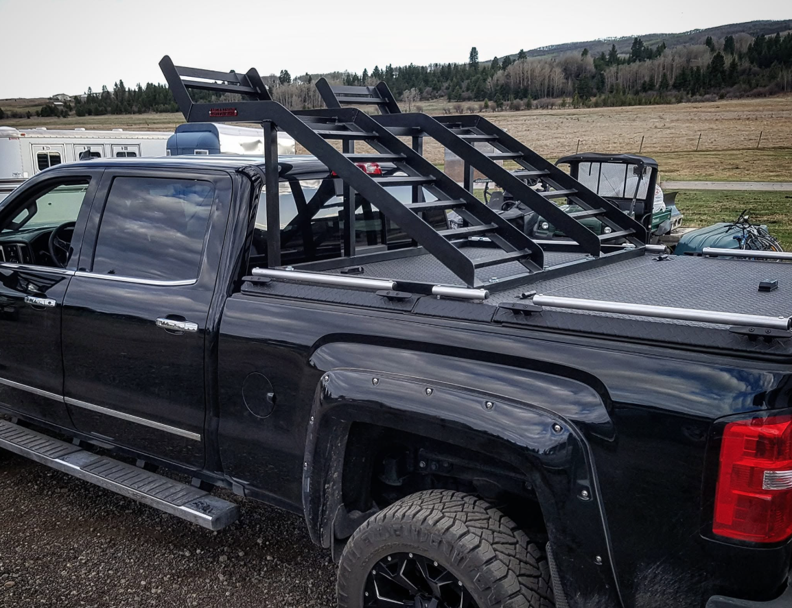 Black pickup truck with ladder racks mounted on its bed, parked outdoors with grassy fields and trees in the background.