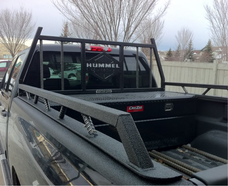 The back of a silver pickup truck with a black metal rack and a tool box in the truck bed, parked outdoors with trees and a fence in the background.
