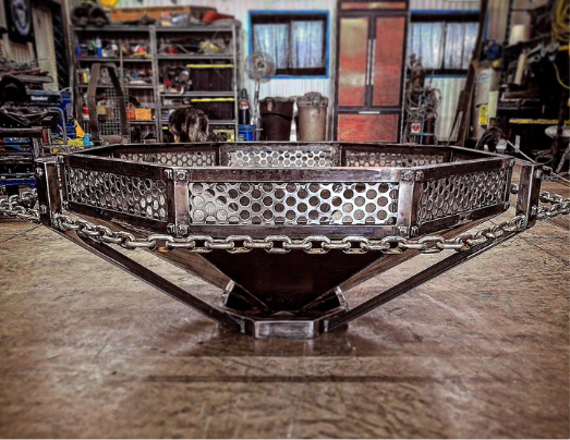 Metal fire pit with perforated sides and chain details, situated on a workshop floor with tools and shelves in the background.