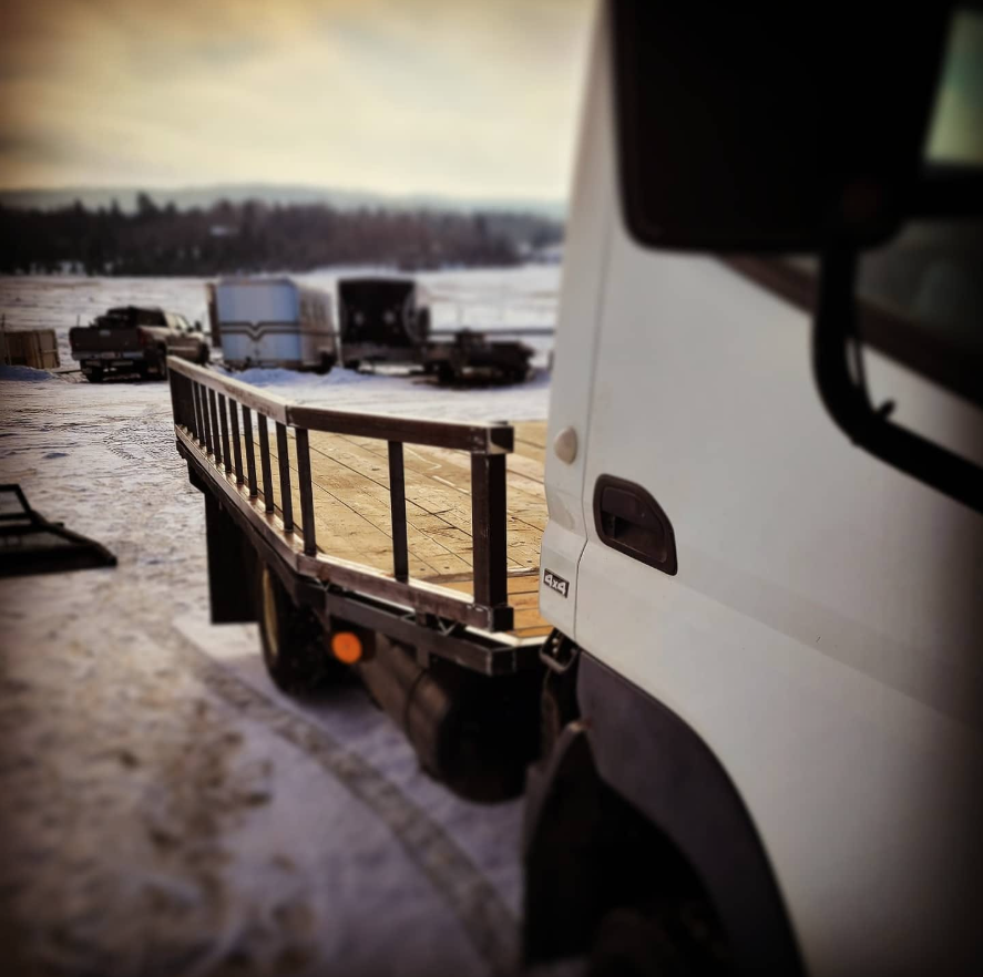 View of a flatbed truck with a wooden deck, parked on snowy ground with other vehicles and trailers in the background and a snow-covered field or lake with trees beyond.