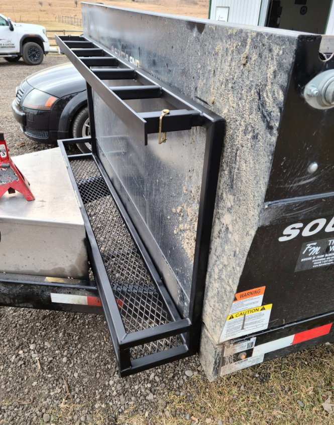A black metal rack attached to a trailer with a textured surface, located outdoors near a black vehicle and other equipment, with warning labels on the trailer.