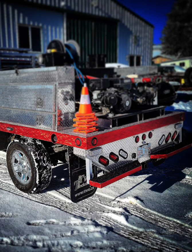 Flatbed trailer with diamond plate metal surface, orange traffic cone, and equipment. Snow on ground and building in background.