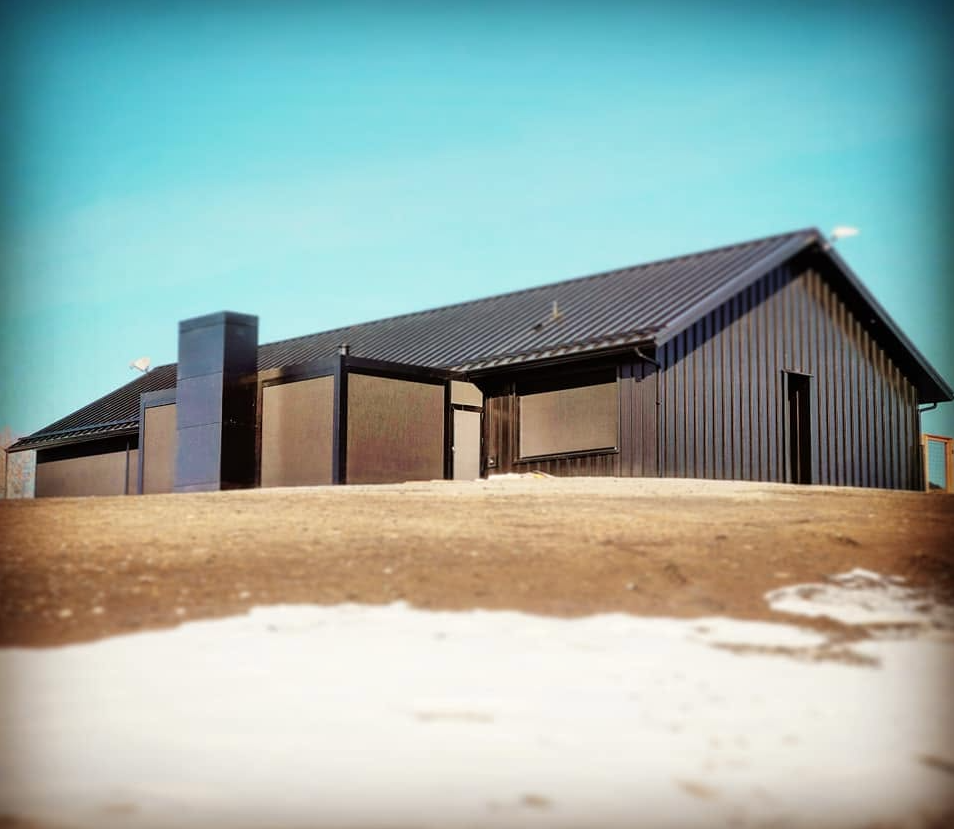 Modern house with dark exterior, pitched roof, and large windows, set on a slightly elevated bare landscape with patches of snow.
