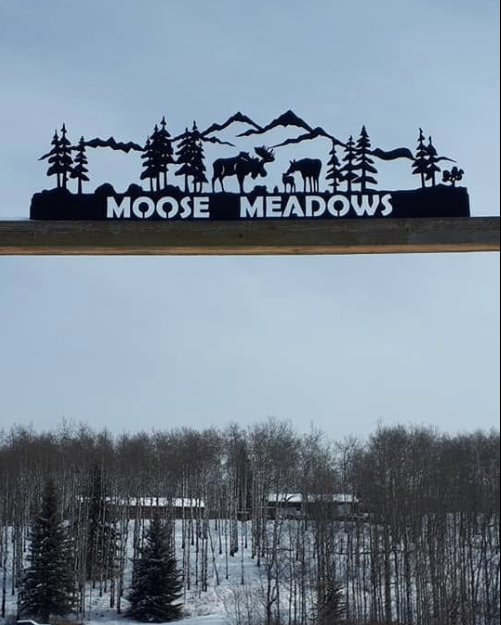 Sign for Moose Meadows featuring mountain, trees, moose, and other wildlife silhouettes against a sky, with snow-covered trees and landscape below.