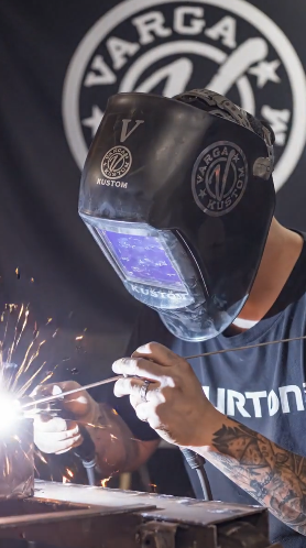 Person welding, wearing a protective welding helmet with the word "VARIGA" and a logo, sparks flying from the welding process.