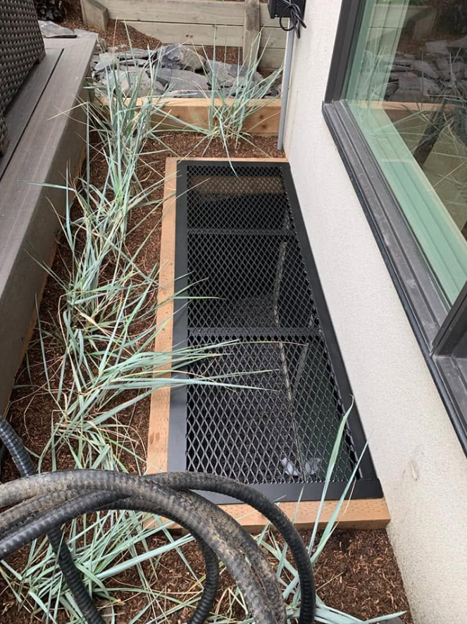 A rectangular black metal grate on a wooden frame beside house wall with a window, with plants and soil around it.