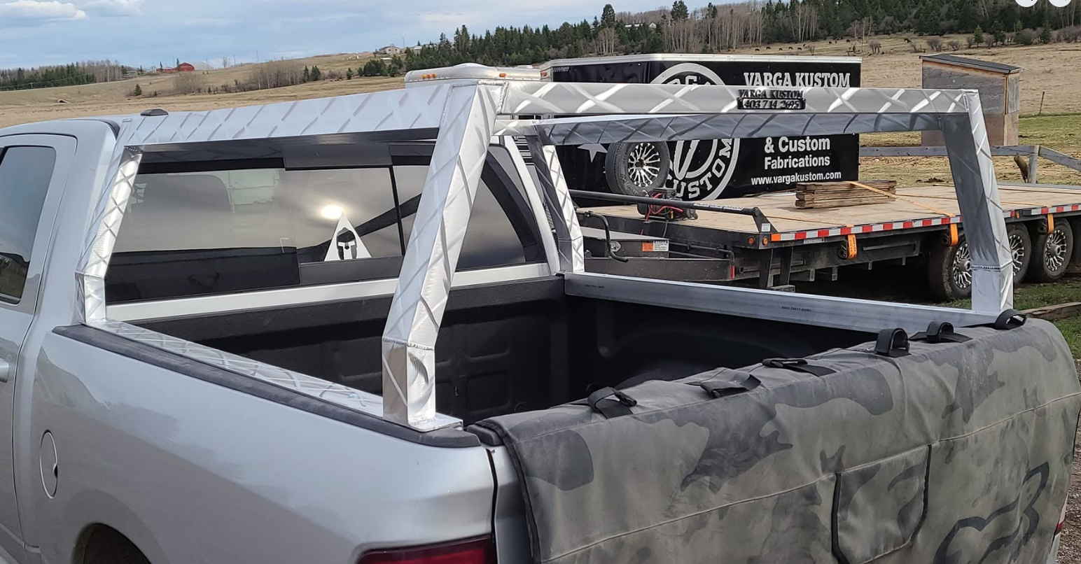 A pickup truck with a custom metal frame installed in the truck bed, near a flatbed trailer carrying a black enclosed trailer with 'Varga Kustom & Custom Fabrications' branding, in a rural outdoor setting with open fields and a partly cloudy sky.