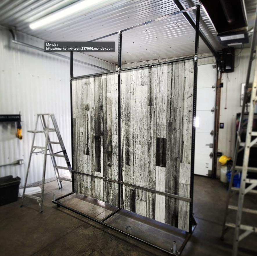 A partially assembled metal frame structure with wood plank panels in a garage or workshop setting, with ladders, tools, and equipment around.