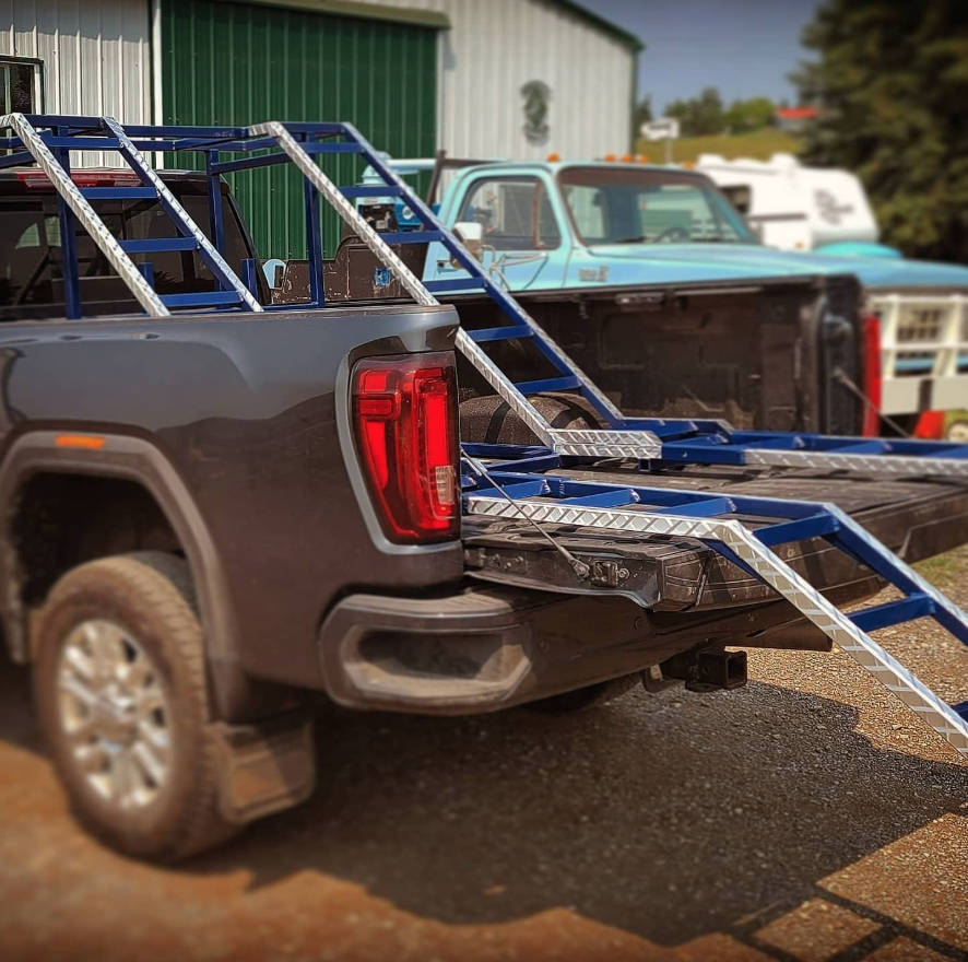 A black pickup truck with a slide-in truck cap, parked outdoors with ladder racks, alongside other trucks and a green building in the background.