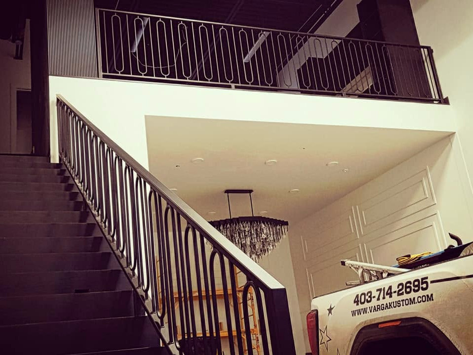 Interior view of a modern building showcasing a staircase with black metal railing, a white wall, and an elegant chandelier hanging from the ceiling. Part of a white vehicle with advertising and a contact number is visible at the bottom right.