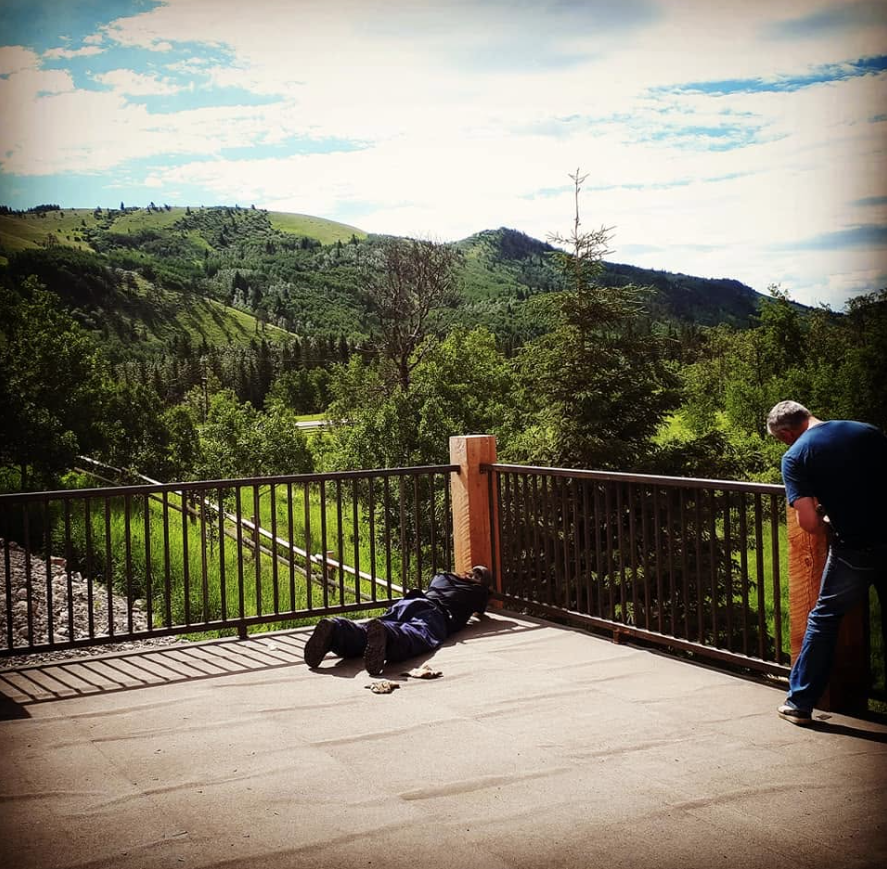 Two men on a balcony overlooking a lush green valley and mountains. One man is lying on the ground, and the other is standing and looking at something over the railing.