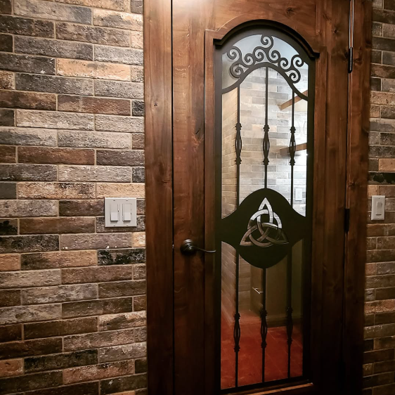 A wooden door with a decorative glass panel featuring a Celtic knot design and a triquetra symbol, set in a brick wall with light switches nearby.