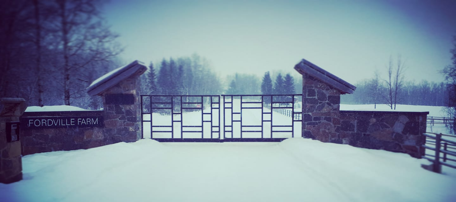 Entrance gate to Fordville Farm covered in snow, with stone pillars and a decorative metal gate, surrounded by snow-covered trees.