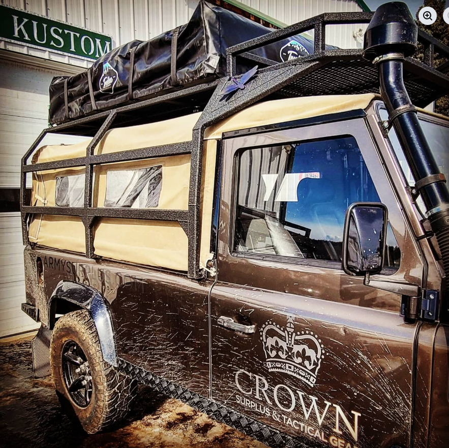 A brown off-road vehicle with a custom rack on top carrying a large cargo bag and a blue inflatable boat, parked inside a garage. The vehicle has branding that reads "CROWN SURPLUS & TACTICAL GEAR" with a crown logo.