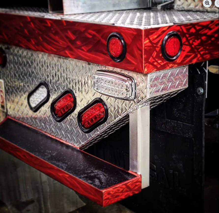 Close-up of the rear corner of a fire truck with red and silver reflective surfaces, circular red tail lights, and rectangular white turn signal lights.