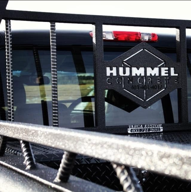 Close-up of the back of a utility truck with a logo for Hummel Concrete, featuring a black diamond shape with white text, and a small sticker with contact information reading 'Varga Kustom' and a phone number.