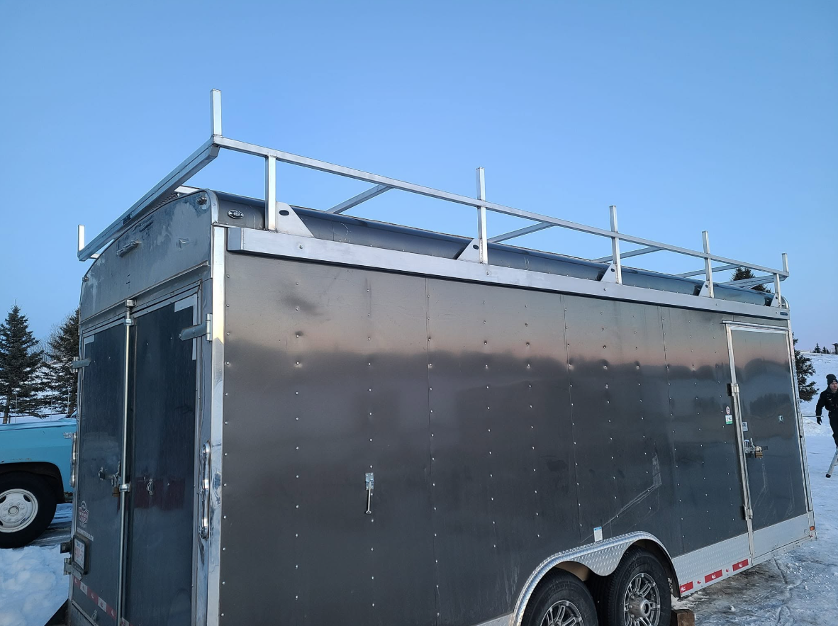 Black enclosed trailer with a metal roof rack, parked on snow with a blue vehicle and trees in the background.