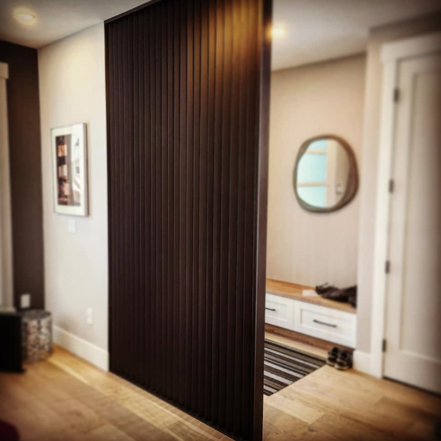 Interior view of a home with a wooden slatted room divider, a small entryway with white drawers, a round mirror, and a door.