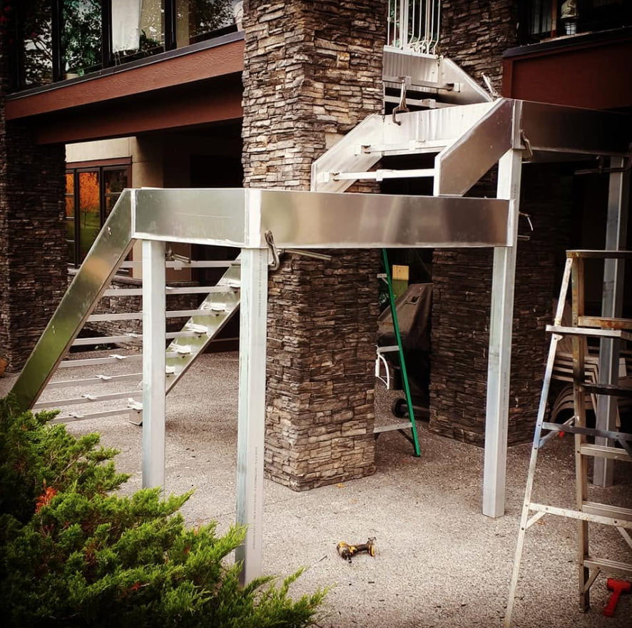 Construction of an outdoor metal staircase with a slide beside a stone pillar and a building with large windows.