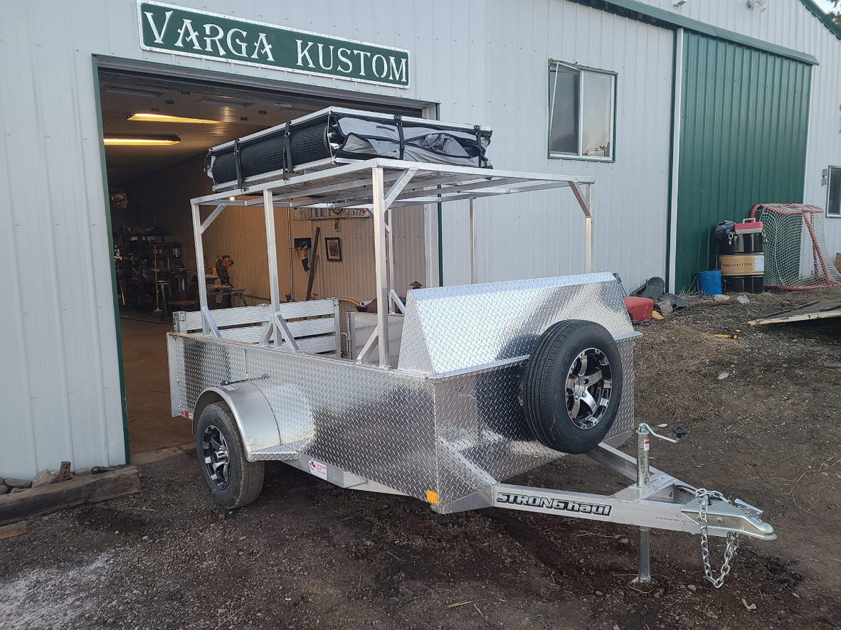 A metal utility trailer parked outside a workshop named Varga Kustom, with an awning and some equipment on top.