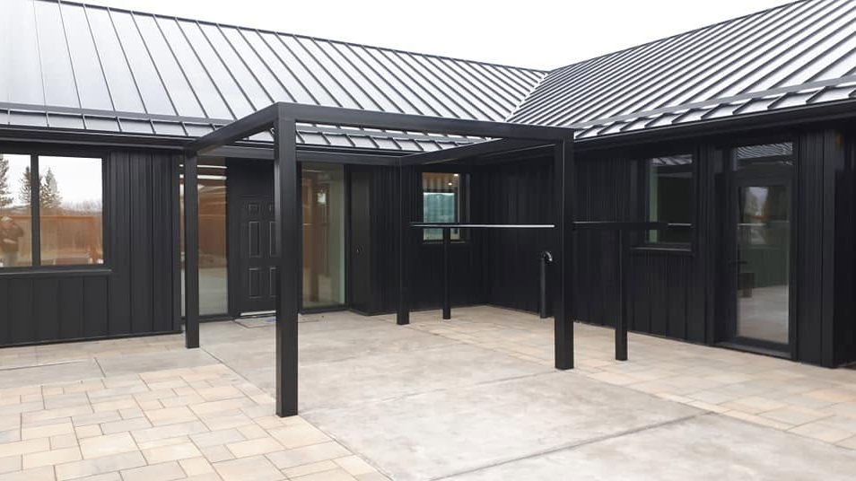 Modern house exterior with black metal siding and large windows, concrete patio, and a covered entrance area.