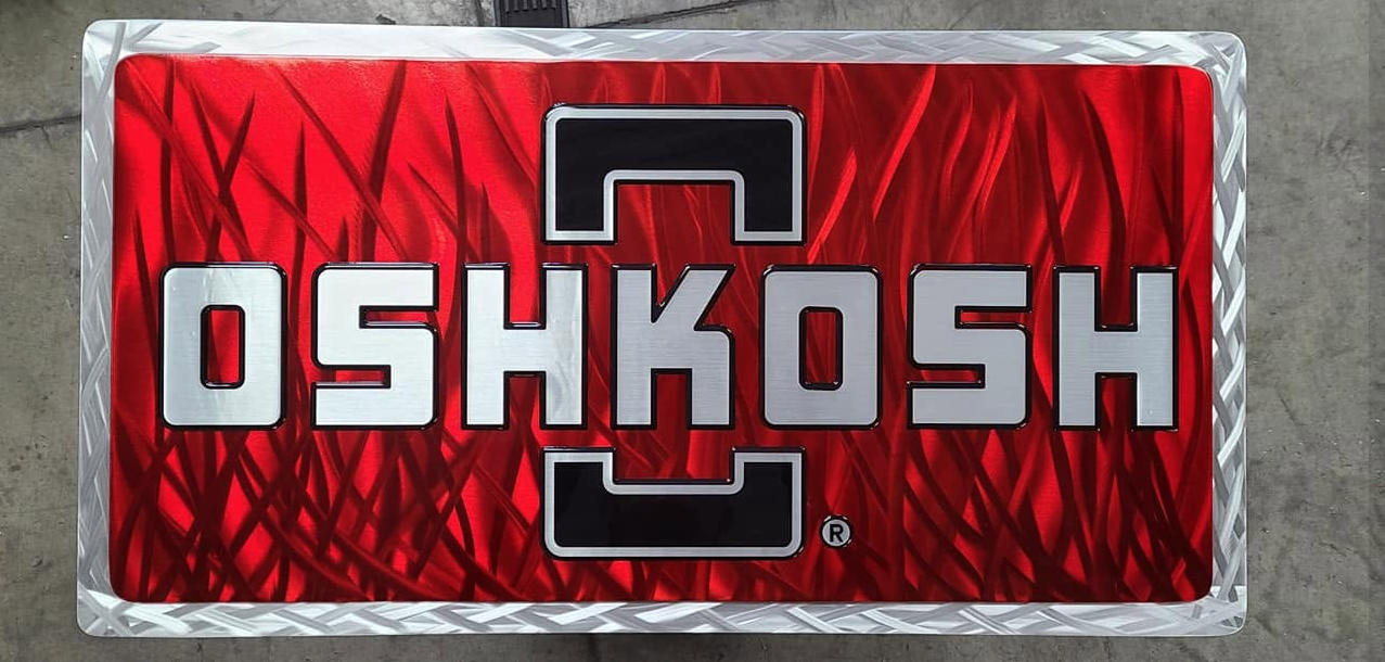 A rectangular sign with a red background and the word 'OSHKOSH' in large white and black letters. The sign has a metallic silver border with a diamond plate pattern.