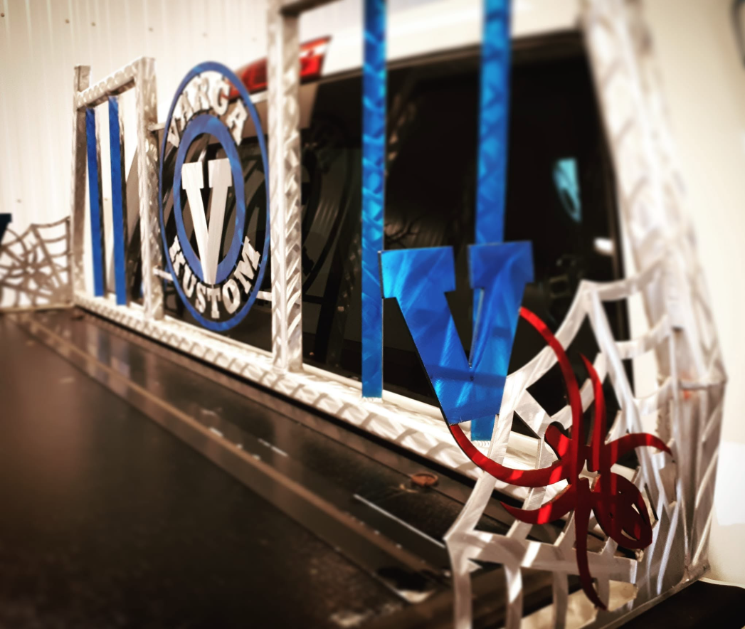 Decorative display with a large letter 'V' in blue, surrounded by letter 'W' in blue, and 'L' in blue, set in a geometric metal framework with patriotic red, white, and blue elements near a vehicle.