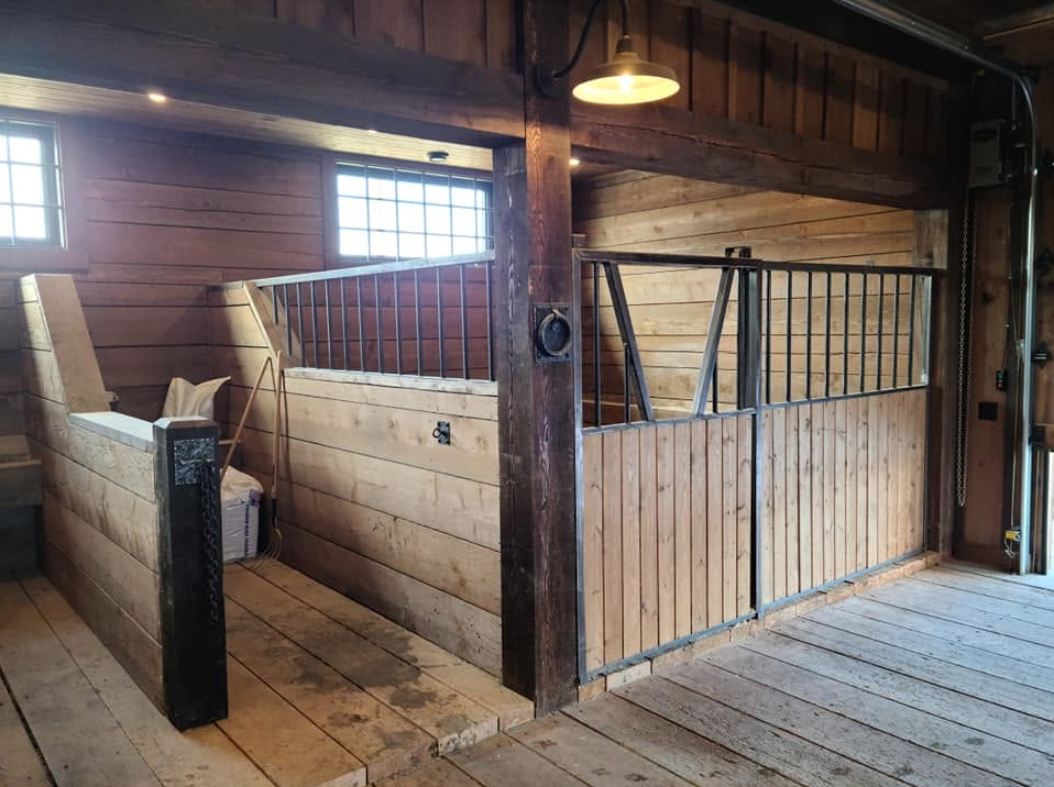 Interior of a barn with two wooden animal stalls, one on the left with a pillow and bedding, and one on the right, both with metal railings, wood-paneled walls, and a wooden floor.