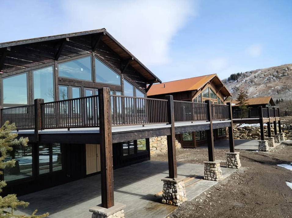Multiple modern houses with large glass windows and wooden balconies in a mountainous area, with a partly cloudy sky and some patches of snow on the ground.