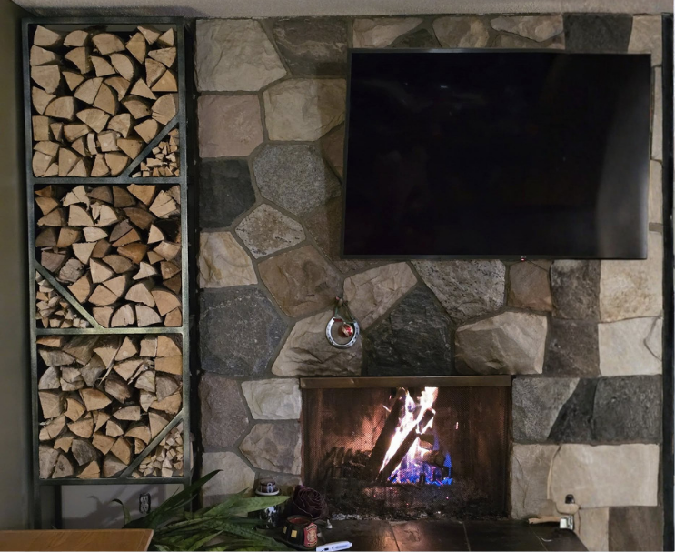 Fire burning in a stone fireplace with a mounted television and a wood storage rack on the side.