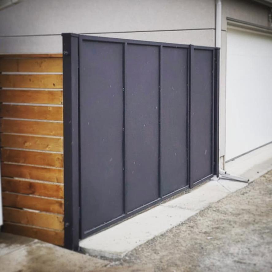 A black metal sliding gate with four vertical panels, installed next to a wooden fence on the left and a gray building wall on the right.