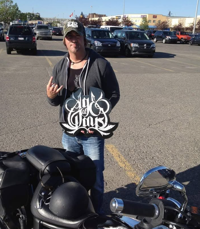 A person standing in a parking lot holding a sign that says "Age of Days" with their left hand making a rock sign gesture. They are wearing a black cap, dark jacket, and jeans. The parking lot is filled with cars, and buildings are visible in the bac