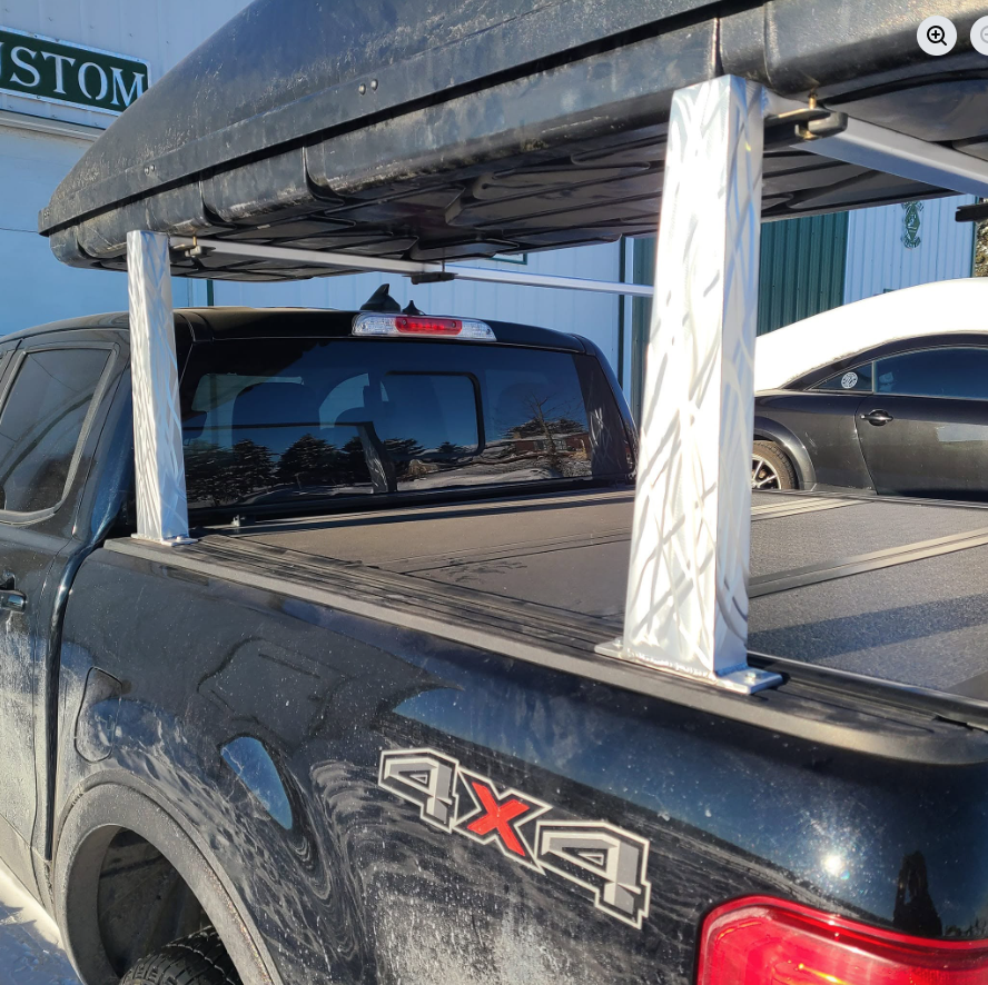 A black pickup truck with a large, covered cargo rack on the bed, equipped with a surfboard underneath, parked outdoors near other vehicles and a building.