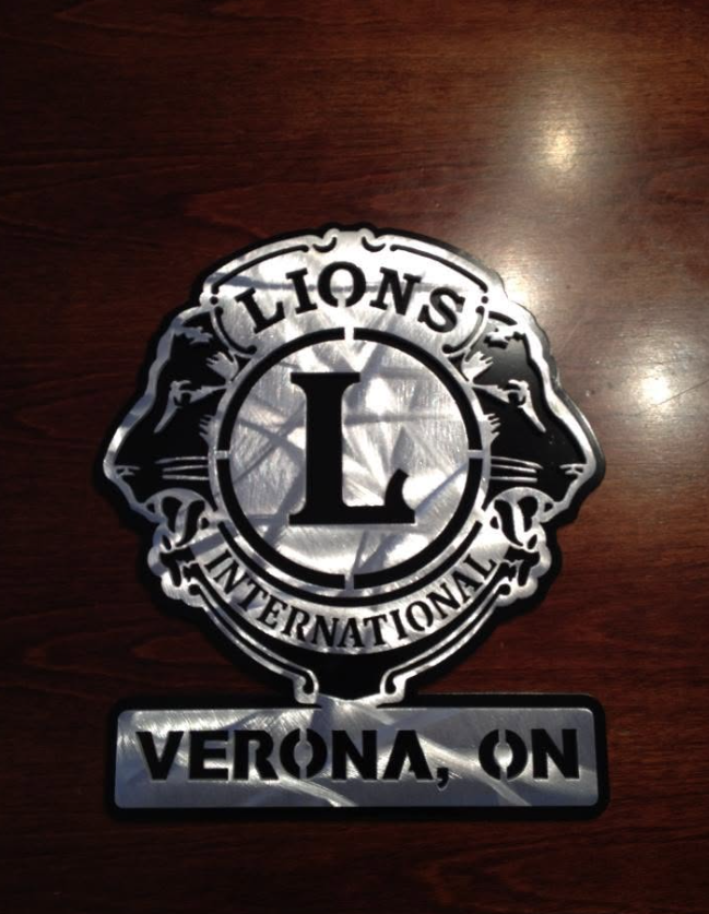 Metal emblem of Lions Clubs International with the location Verona, ON engraved below, placed on a wooden surface.