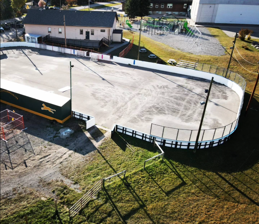 An aerial view of an outdoor ice skating rink surrounded by a black and white barrier, with a small sports field and playground nearby in a residential area.