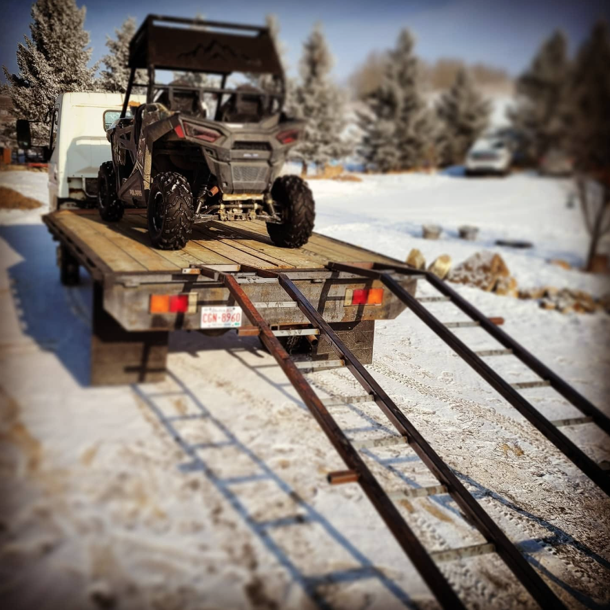 A flatbed truck with a UTV on it, with ramps extended down to the snowy ground for loading or unloading, in a winter landscape with snow-covered trees and a car in the background.