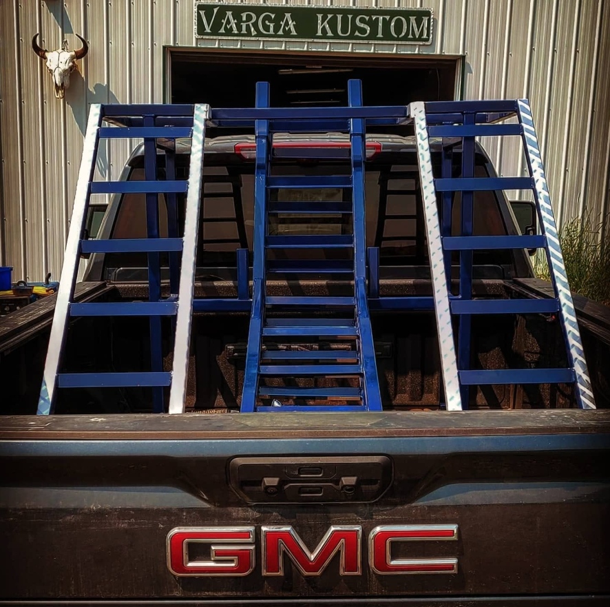 A GMC pickup truck with three blue and white ladders in its bed, parked outside a building labeled 'Varga Kustom.' A bull skull with horns hangs on the pole wall to the left of the building.