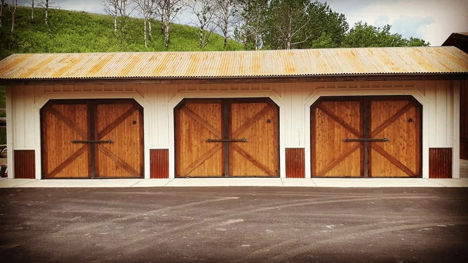 Three wooden garage doors on a barn with a metal roof, set against a background of trees and a hillside.