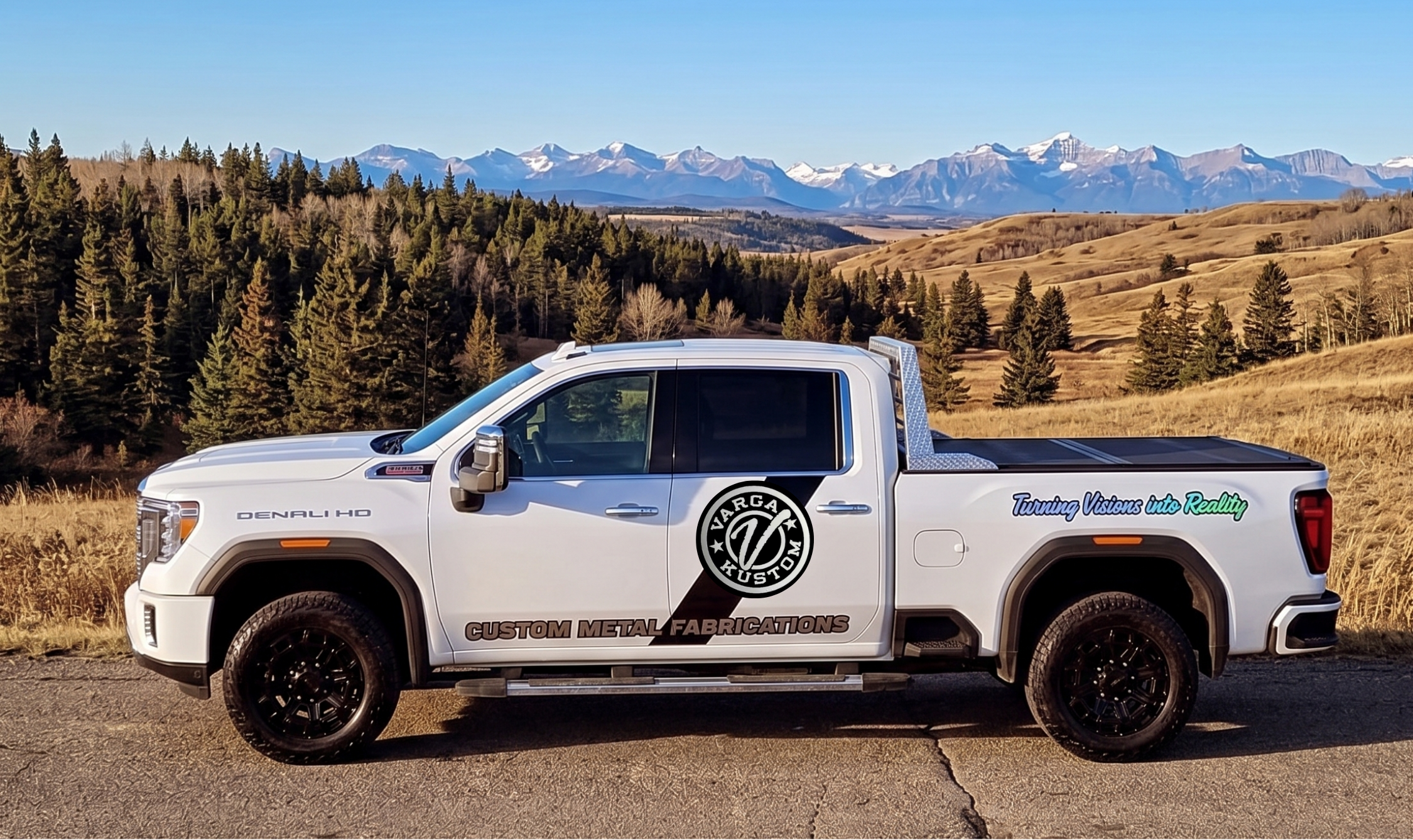 White pickup truck with custom logos and text on its side parked on a rural road, with a backdrop of trees, rolling hills, and snow-capped mountains under a clear blue sky.