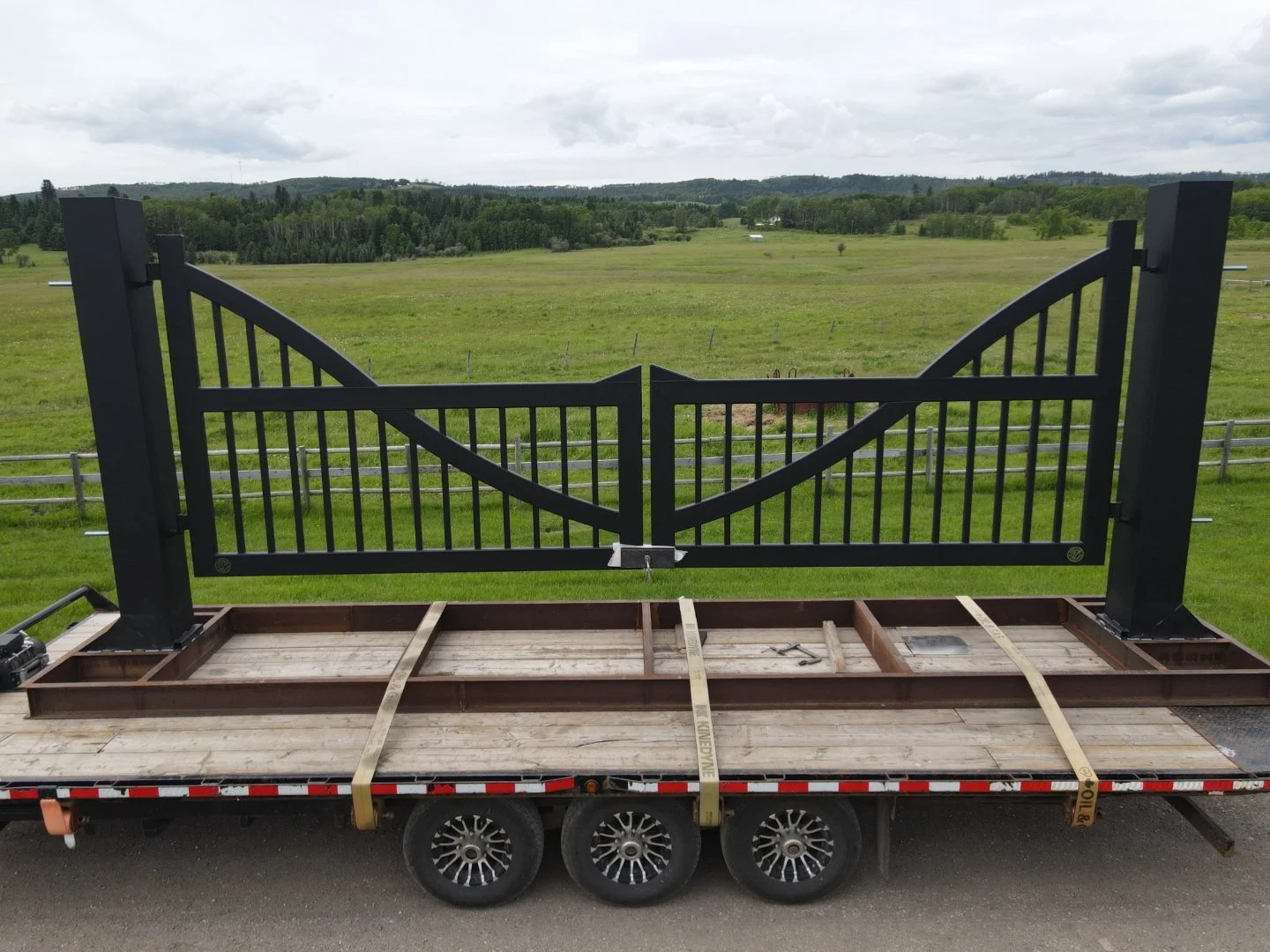 A black metal gate with a decorative curve is mounted on a wooden frame on a flatbed trailer, overlooking a green countryside landscape with fields, trees, and a cloudy sky.