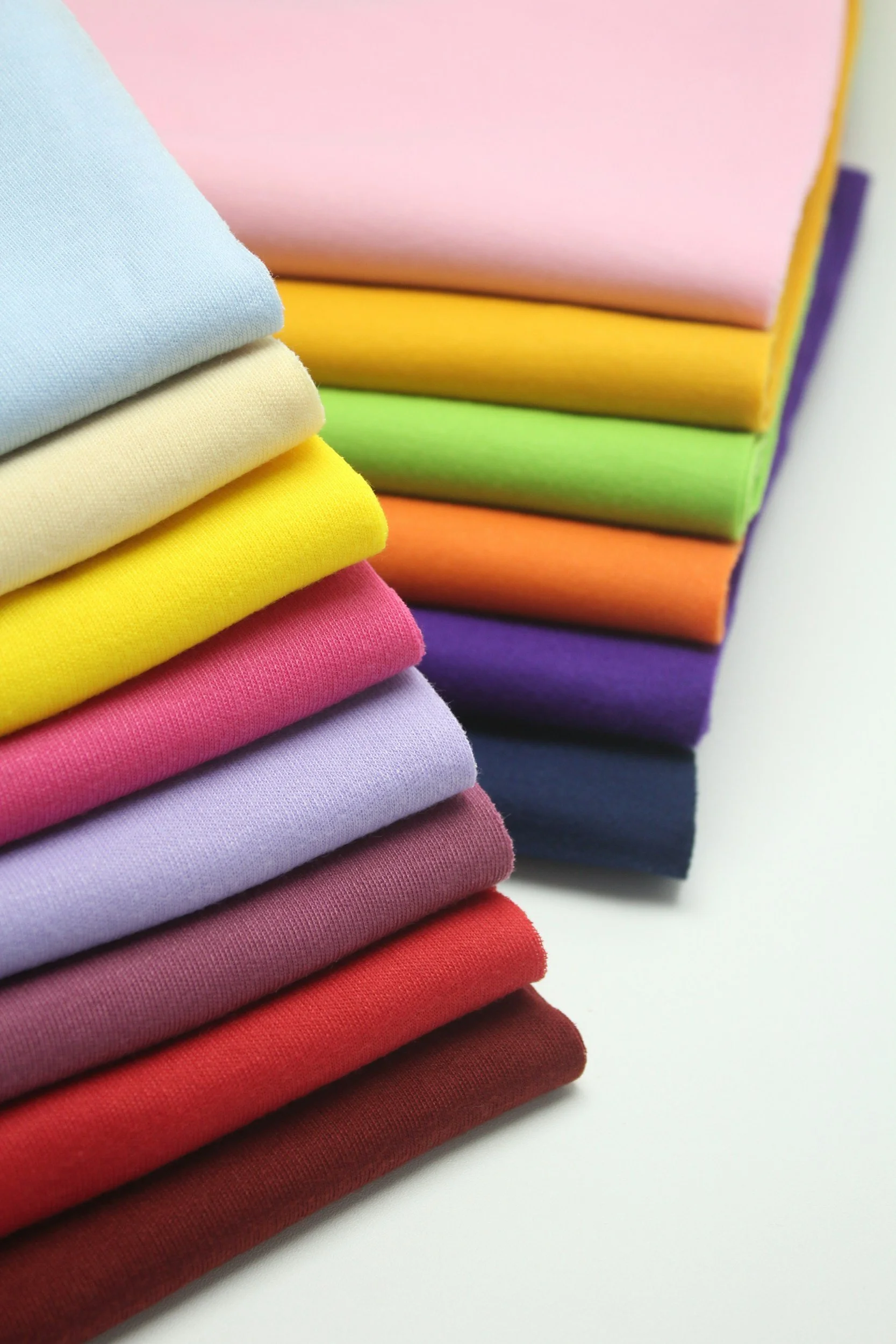 Stacked colorful folded fabrics arranged in rainbow order from top to bottom: light blue, pale yellow, yellow, green, orange, purple, pink, lavender, red, and maroon.