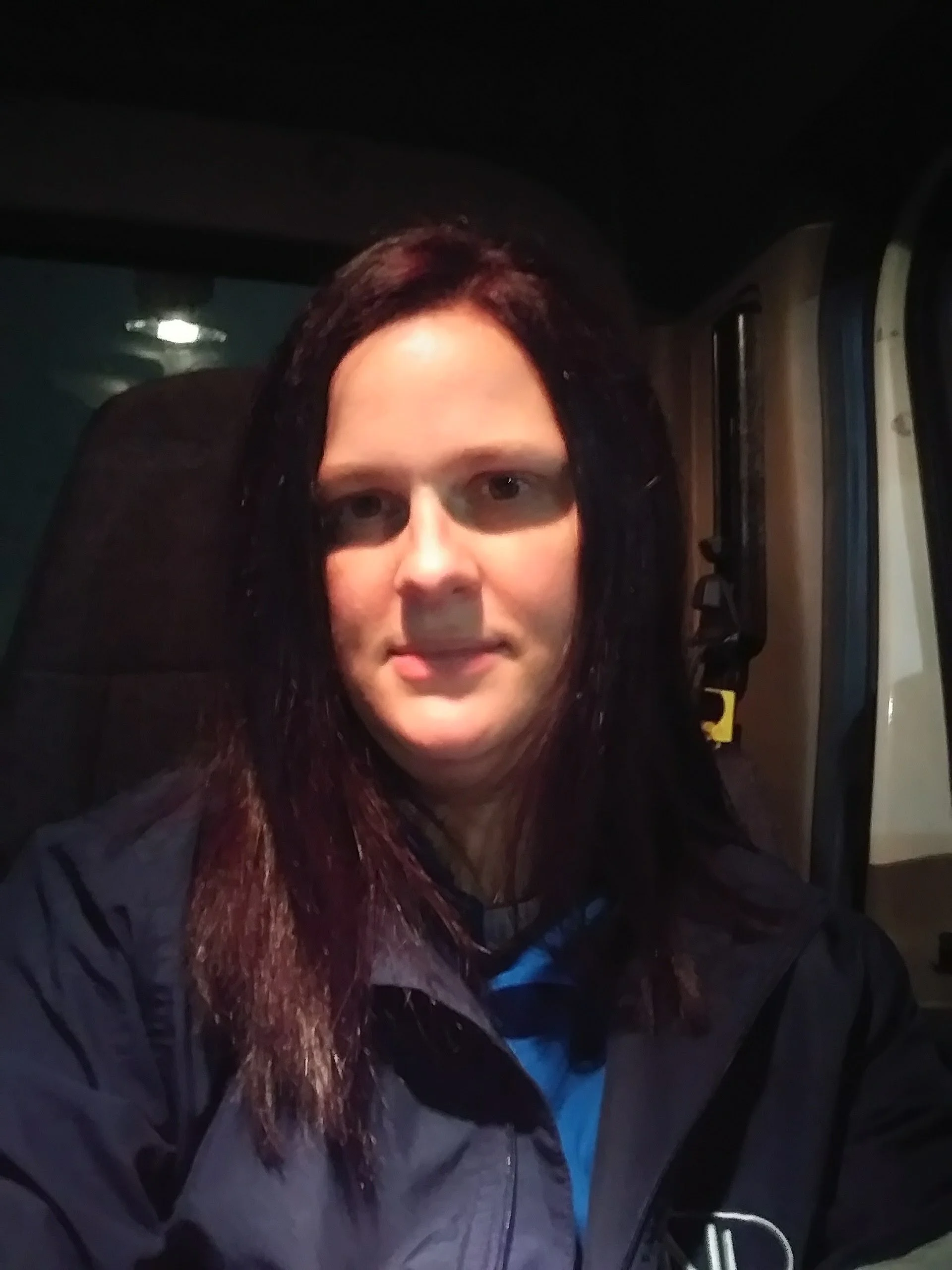 Close-up of a person with long dark hair, sitting inside a vehicle, looking at the camera, with a seatbelt across the chest, at night with dim lighting.