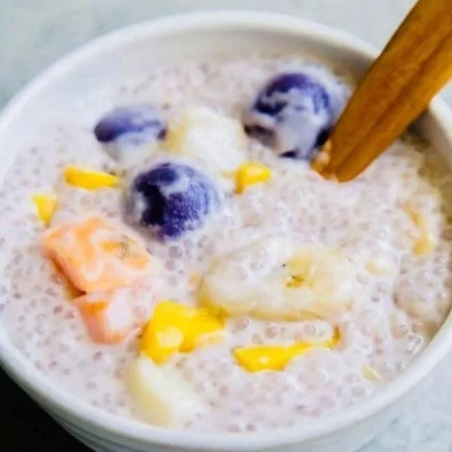 A bowl of mixed fruit with blueberries, bananas, and mango chunks in a creamy liquid, with a wooden spoon inside.