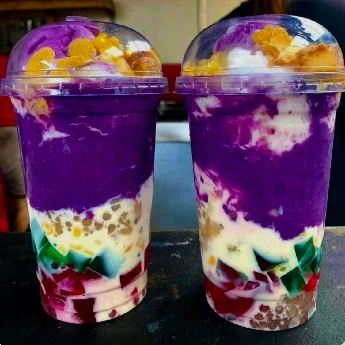 Two colorful layered drinks in clear plastic cups with dome lids, topped with cereal and possibly fruit, featuring purple, white, yellow, and red layers.