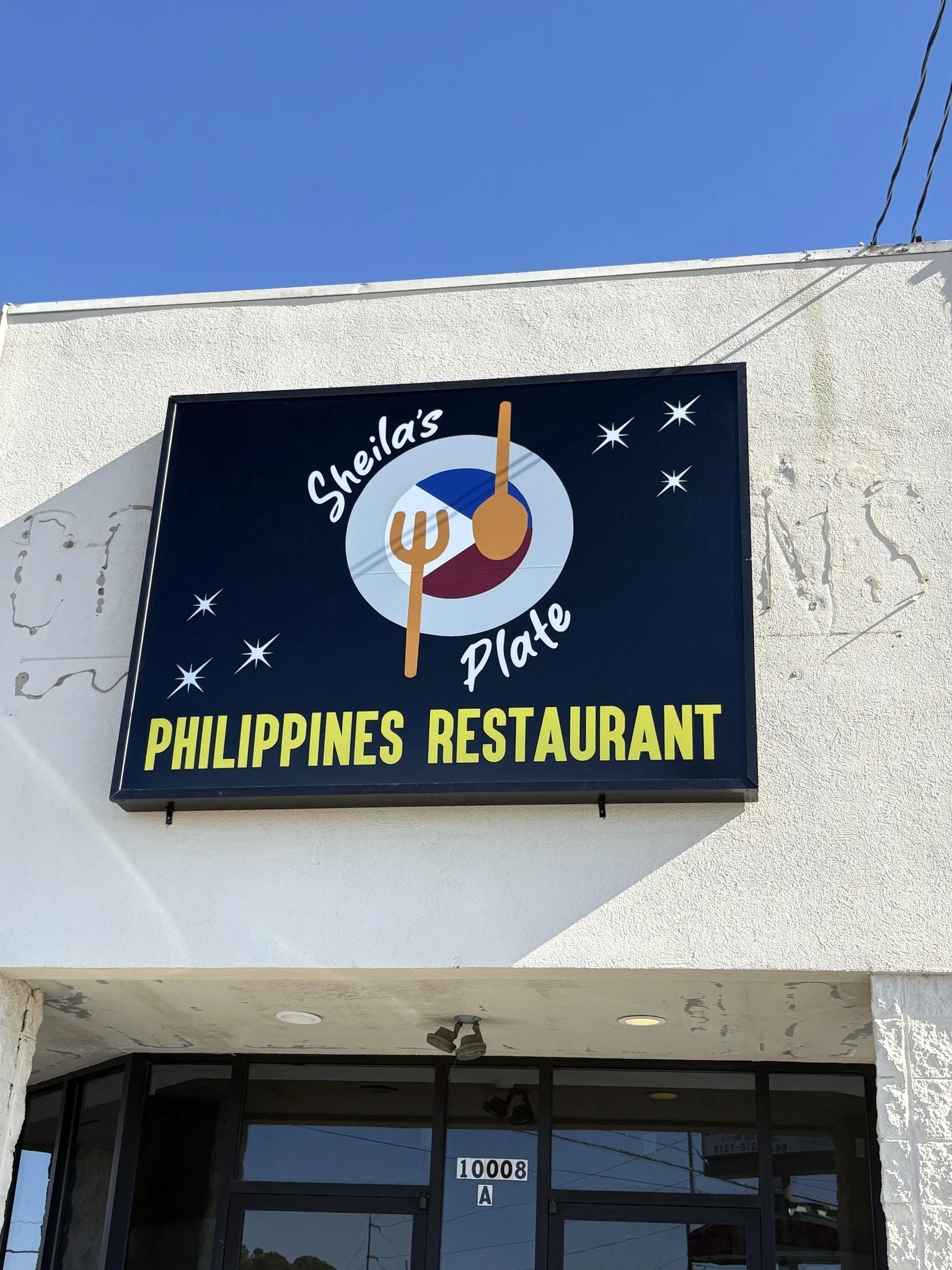 Signboard for Sheila's Plate Restaurant with logo featuring a spoon and fork on a circular background, and stars around it, on a white wall outside the building.